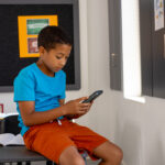 In school, in a classroom, a young biracial boy is sitting, focusing on a smartphone. He has short black hair, wearing a blue shirt and orange shorts, unaltered.